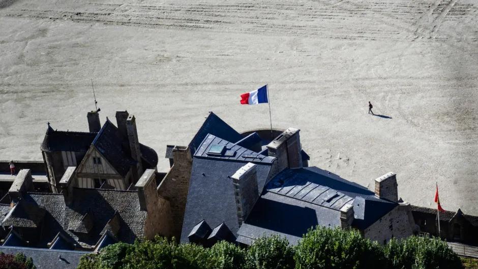 Blick von oben auf Gebäude der Abtei Mont-Saint-Michel mit französischer Flagge und Sandflächen im Hintergrund, Normandie, Frankreich.