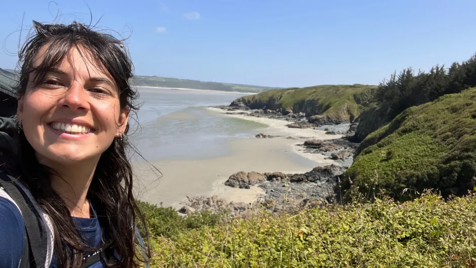 Unsere Fahrradreise-Expertin Johanne macht ein Selfie an der bretonischen Küste. Im Hintergrund sieht man eine ruhige Bucht mit Sandstrand, Felsen und grünen Klippen unter blauem Himmel.