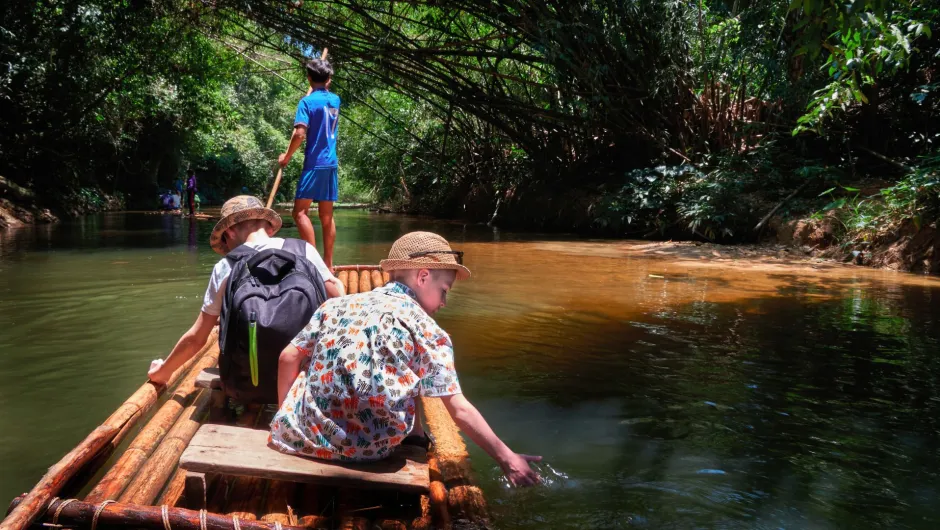 Zwei Kinder sitzen auf einem Bambusfloß auf einem ruhigen Fluss im dichten Regenwald, während ein Mann das Floß mit einer Stange steuert.