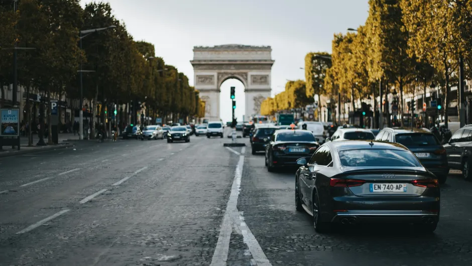 Autos fahren auf einer breiten, von Bäumen gesäumten Straße in Paris, im Hintergrund ist der Arc de Triomphe zu sehen.