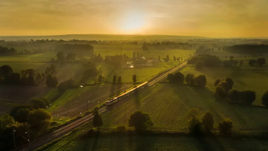 Zug fährt bei Sonnenaufgang durch eine ländliche Landschaft mit Feldern, Bäumen und leichtem Morgennebel.