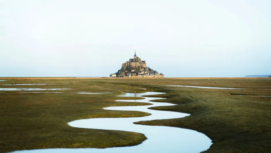 Mont-Saint-Michel in der Normandie mit geschwungenem Wasserlauf im Watt bei Ebbe und Blick auf die Abtei in der Ferne.