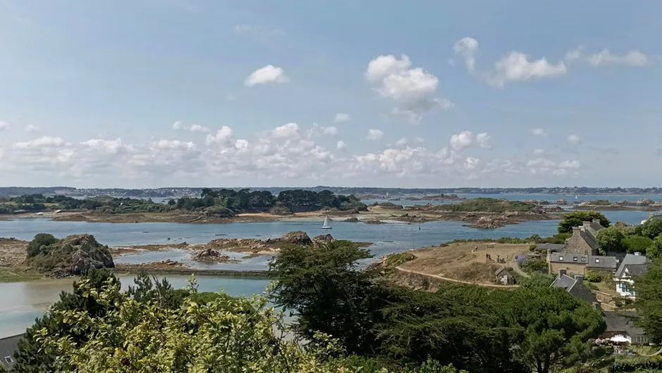 Weite Küstenlandschaft mit kleinen Inseln und ruhigem Wasser, ein Segelboot in der Mitte, im Vordergrund Bäume und rechts einige Häuser unter einem Himmel mit lockeren Wolken.