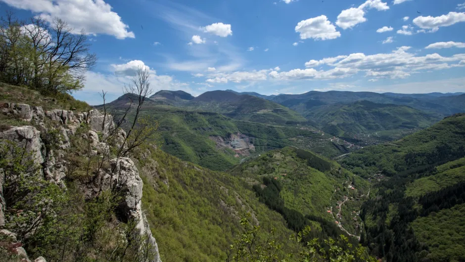 Grüne Berglandschaft mit felsigem Aussichtspunkt, bewaldeten Tälern und weitem Blick unter blauem Himmel mit Wolken.