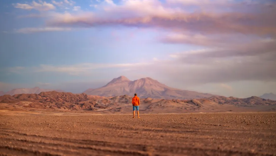 A man stadning in the middle of the Atacama Desert in Chile. Behind him is a mountain out of the distinct red stone found in this desert.