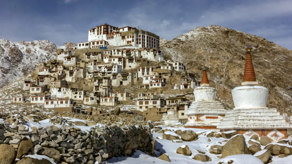 Schneebedecktes Bergdorf in Ladakh, Indien, mit weißem Kloster auf einem Hügel und buddhistischen Stupas im Vordergrund.