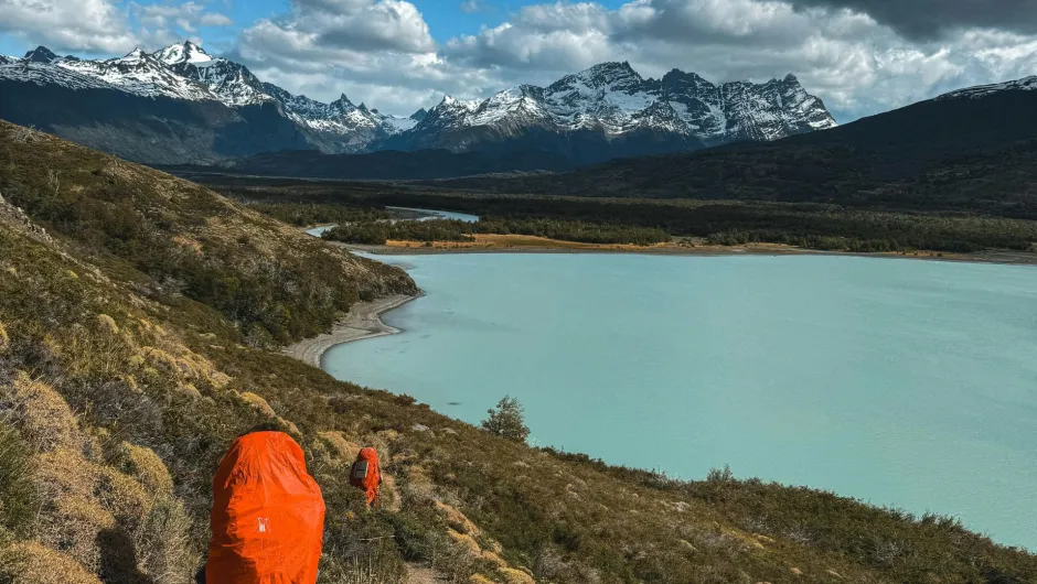Wanderer mit orangefarbenen Rucksackhüllen auf einem schmalen Pfad oberhalb eines türkisfarbenen Sees, umgeben von schneebedeckten Bergen und dramatischen Wolken.