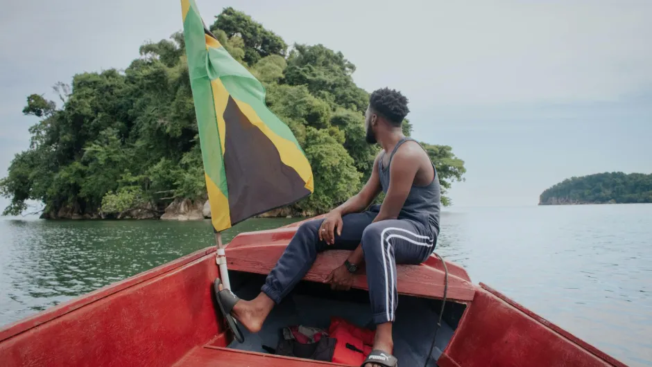 Ein Mann sitzt vorne in einem roten Boot auf ruhigem Wasser, neben einer grün-gelb-schwarzen Flagge, mit bewachsenen Inseln im Hintergrund.