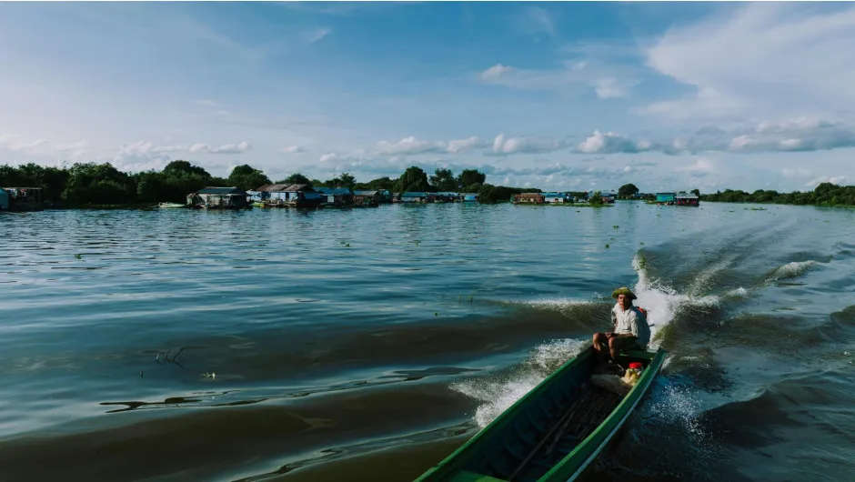 Ein Schnellboot fährt über einen weiten See in Kambodscha, vorbei an schwimmenden Häusern unter blauem Himmel.