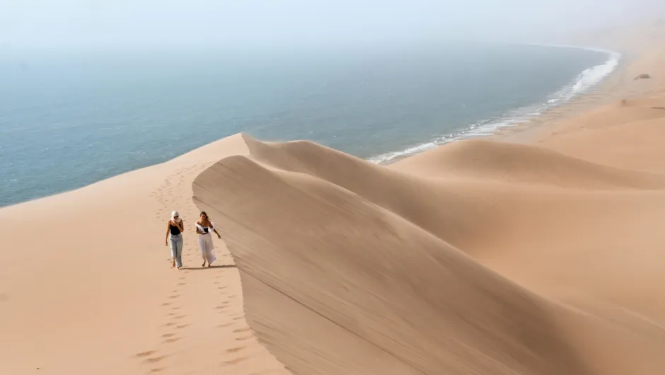 Zwei Personen laufen über hohe Sanddünen an einer nebligen Küste, wo die Wüste direkt auf den Ozean trifft.