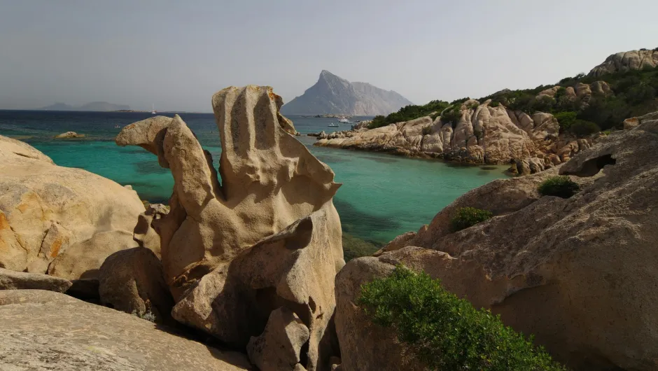 Felsige Küste auf Sardinien mit türkisfarbenem Wasser, bizarren Granitfelsen und einer Berginsel im Hintergrund.