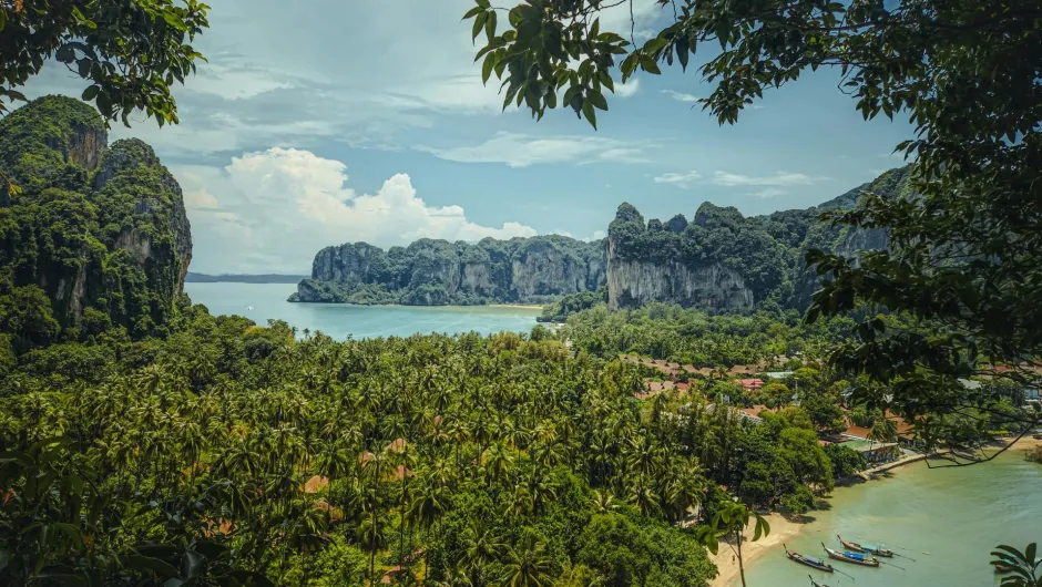 Panoramablick auf eine tropische Bucht in Thailand mit türkisfarbenem Wasser, Longtail-Booten, dichtem Palmenwald und steilen, bewachsenen Kalksteinfelsen unter blauem Himmel.
