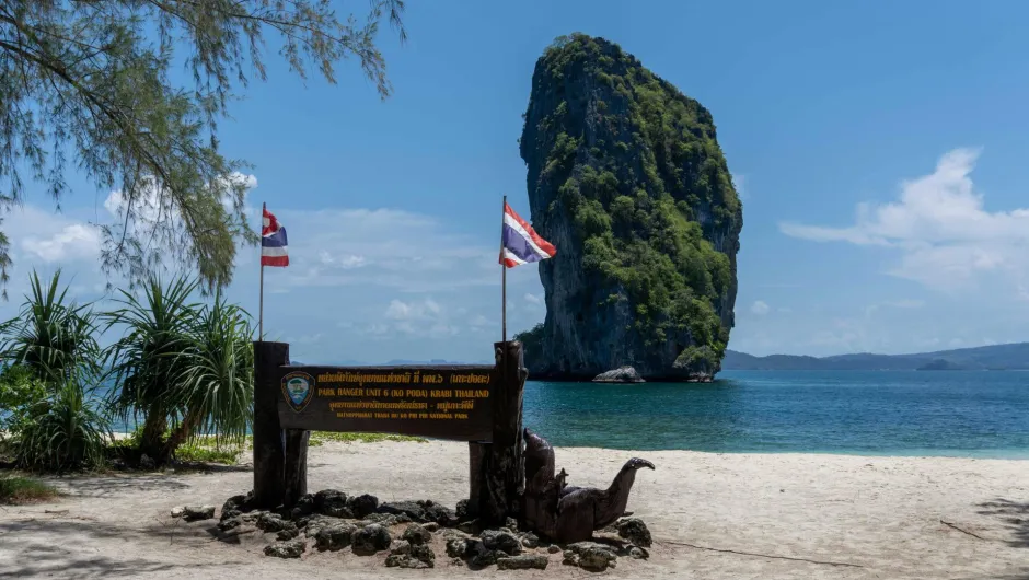 Tropischer Sandstrand in Thailand mit zwei thailändischen Flaggen, einem Nationalpark-Schild und einem hohen, bewachsenen Kalksteinfelsen im türkisblauen Meer unter klarem Himmel.
