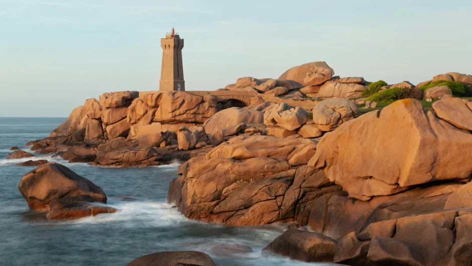 Ein Leuchtturm steht auf einer zerklüfteten rosafarbenen Granitküste am Meer. Die warm beleuchteten Felsen und das sanft bewegte Wasser erzeugen eine ruhige Küstenlandschaft.