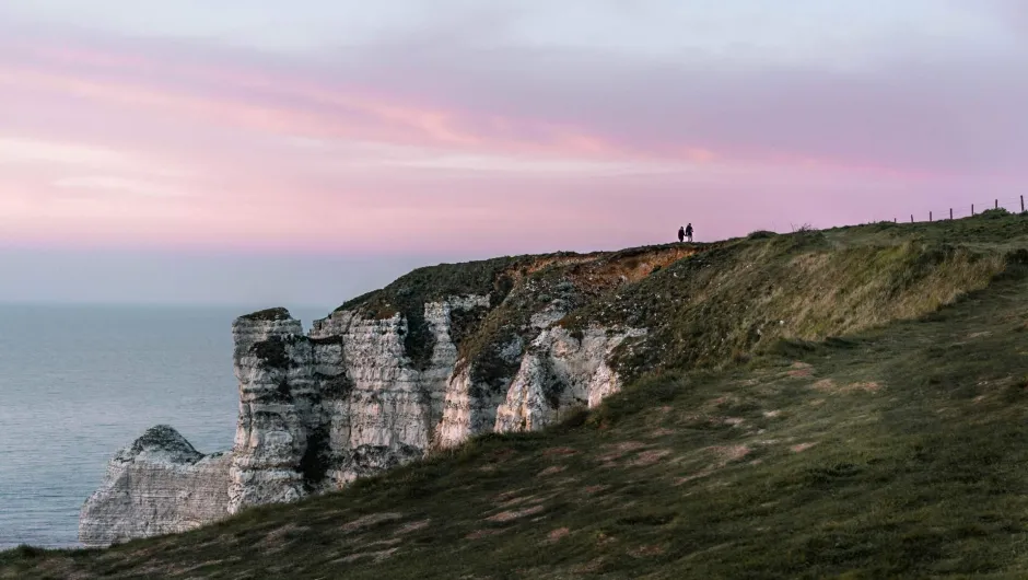 Zwei Personen stehen auf grasbewachsenen Kreidefelsen an der Küste, während der Himmel bei Sonnenuntergang rosa und violett gefärbt ist.