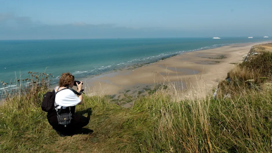 Eine Person kniet mit einer Kamera auf einer grasbewachsenen Klippe und blickt auf einen weiten Sandstrand und das blaue Meer mit Schiffen am Horizont.