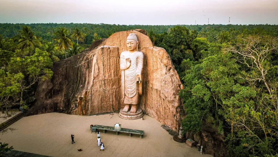 Eine große, in Fels gehauene Buddha-Statue steht inmitten einer grünen Waldlandschaft; davor befinden sich einige Besucher auf einem offenen Platz.