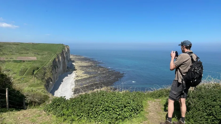 Ein Wanderer mit Rucksack steht auf einer grünen Küstenklippe und fotografiert die steilen Felsen und das Meer. Unterhalb der Klippen erstreckt sich eine felsige Küste, darüber liegt ein klarer blauer Himmel.