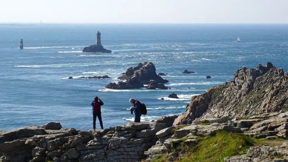 Zwei Wanderer an der felsigen Küste der Bretagne mit Blick auf den Atlantik und mehrere Leuchttürme.