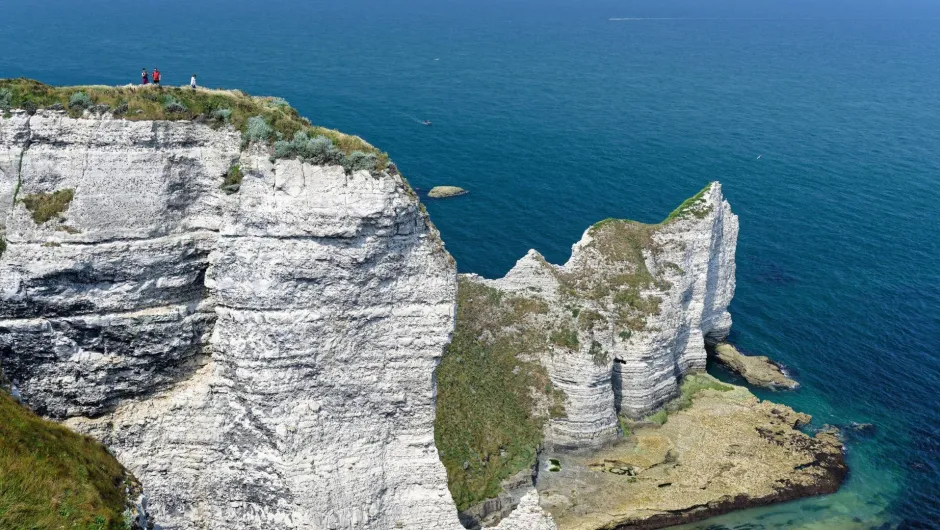 Steile weiße Kreidefelsen an einer tiefblauen Meeresküste. Auf der grasbewachsenen Klippe stehen einige Menschen. Rechts ragt eine schmale, zerklüftete Felsformation ins Meer; am Fuß der Klippen liegen Felsen und ein kleiner Küstenabschnitt.