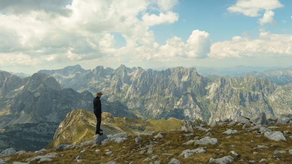Person auf einem Berggipfel mit Blick auf eine weitläufige, zerklüftete Gebirgslandschaft unter blauem Himmel mit Wolken..