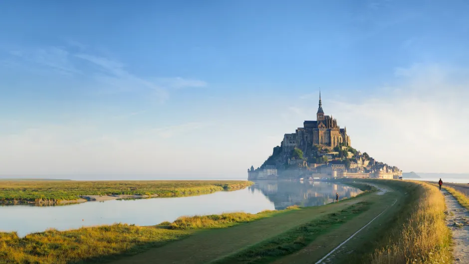 Blick über grüne Salzwiesen und einen schmalen Wasserlauf auf den Mont-Saint-Michel mit seiner hoch aufragenden Abtei im warmen Morgenlicht.
