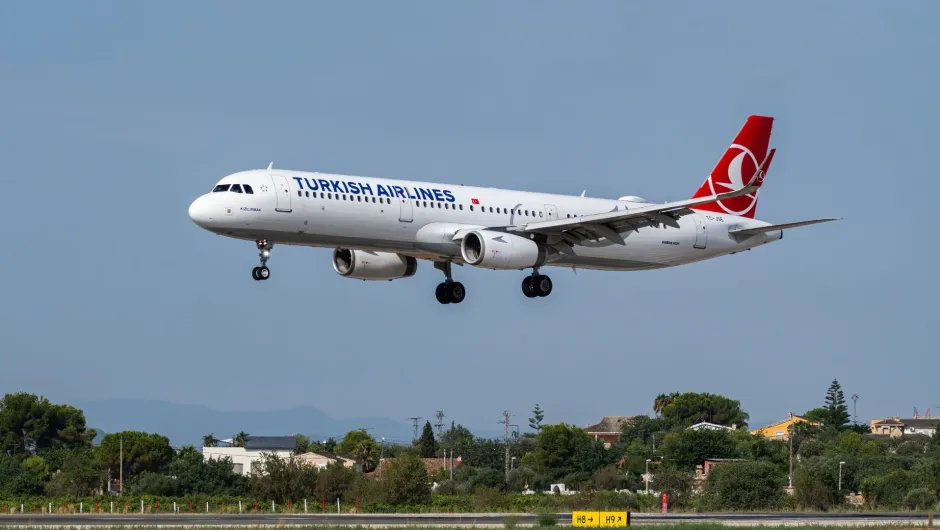 Ein weißes Passagierflugzeug von Turkish Airlines befindet sich mit ausgefahrenem Fahrwerk im Landeanflug. Im Hintergrund sind ein blauer Himmel sowie Bäume und Häuser zu sehen.