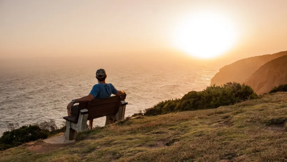 Ein Mann sitzt auf einer Bank und blickt bei Sonnenuntergang auf den Ozean, was eine ruhige Szene schafft.