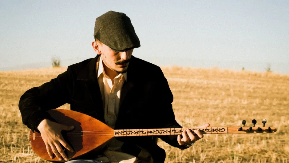Ein Mann mit Schiebermütze sitzt in einem trockenen Feld und spielt ein traditionelles Saiteninstrument.