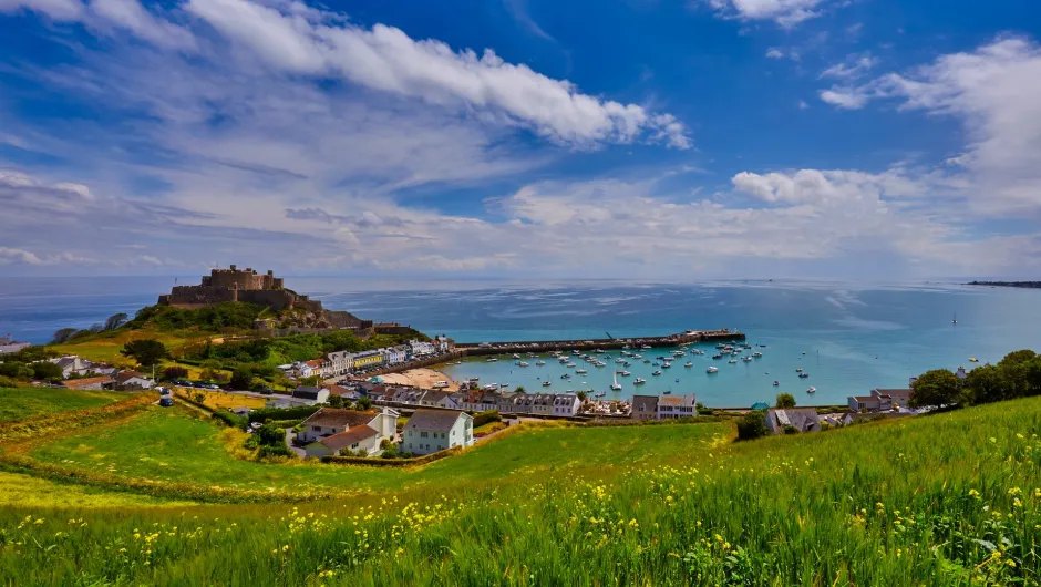 Panoramablick über eine grüne Küstenlandschaft der Kanalinseln mit Hafen, zahlreichen Segelbooten und einer historischen Burg am Meer unter blauem Himmel.