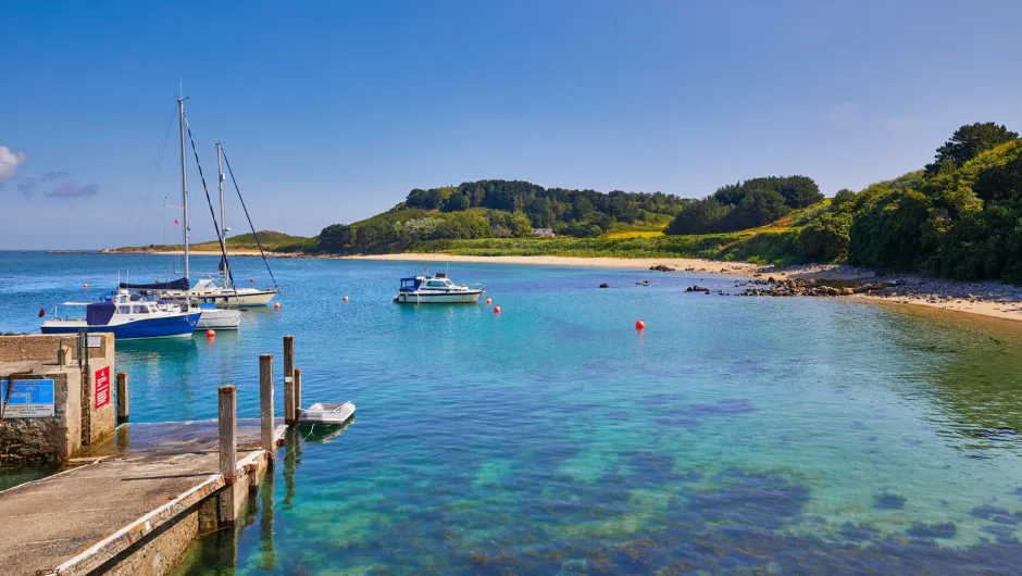 Ruhige Bucht auf Herm mit klarem türkisblauem Wasser, mehreren Segel- und Motorbooten sowie einem kleinen Anleger im Vordergrund. Im Hintergrund liegen ein heller Sandstrand und grüne, dicht bewachsene Hügel unter blauem Himmel.