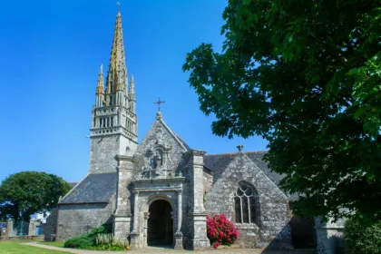 Historische Steinkirche in der Bretagne mit hohem Kirchturm, kunstvoll gestaltetem Eingangsportal und blühenden Pflanzen vor dem Gebäude unter blauem Himmel.