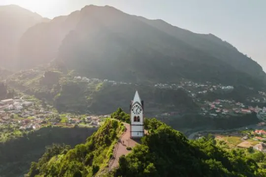 Eine Kirche auf einem grünen Berg, mit leicht grauem, verwaschenem Himmel, durch den links die Sonne scheint.
