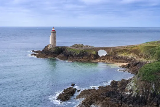 Leuchtturm von Kermorvan an der felsigen Küste der West-Bretagne mit Steinbrücke und Atlantik im Hintergrund.