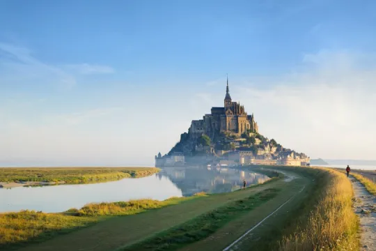 Blick über grüne Salzwiesen und einen schmalen Wasserlauf auf den Mont-Saint-Michel mit seiner hoch aufragenden Abtei im warmen Morgenlicht.