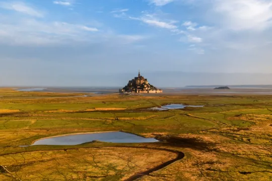 Schafe grasen auf einer weiten grünen Wiese vor dem Mont Saint-Michel, der im Hintergrund unter blauem Himmel aufragt.