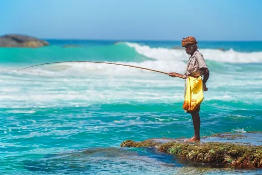 Ein älterer Fischer steht barfuß auf einem flachen Felsen im türkisblauen Meer und hält eine lange, gebogene Angelrute über das Wasser. Hinter ihm brechen weiße Wellen unter einem klaren blauen Himmel.